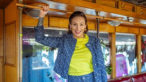 Zu sehen ist Berlin Tag und Nacht Darstellerin Liza Waschke, die in der Serie Milla spielt. Sie steht in einer Art alten Bahn und hält sich an einer Stange oben fest. Sie trägt ein blau gemustertes Anzugset und ein neongrünes Shirt - Foto: RTL II