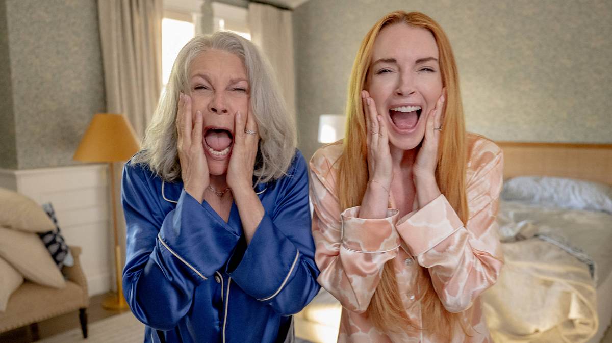 Man sieht Jamie Lee Curtis in einem blauen Pyjama und Lindsay Lohan in einem rosa Pyjama in einem Schlafzimmer stehen. Die beiden schauen entsetzt nach vorne.