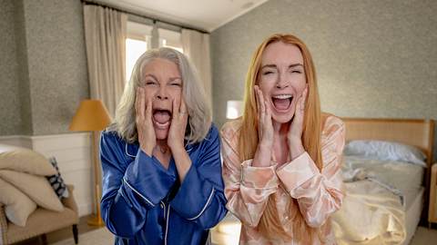 Man sieht Jamie Lee Curtis in einem blauen Pyjama und Lindsay Lohan in einem rosa Pyjama in einem Schlafzimmer stehen. Die beiden schauen entsetzt nach vorne. - Foto: IMAGO / Cinema Publishers Collection
