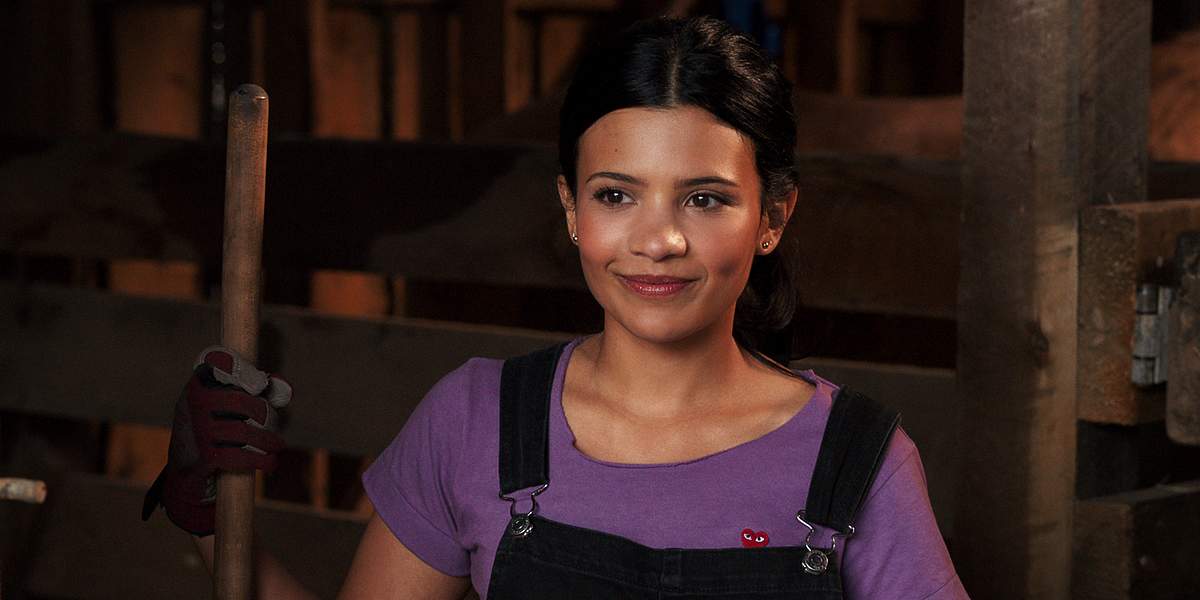 Jackie trägt ein lilanes t-Shirt und einen Overall aus Jeans und lächelt leicht 
