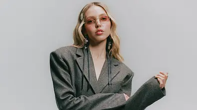 LINA schaut in die Kamera, trägt einen grauen oversized Blazer und eine kleine Brille mit rosagefärbten Gläsern - Foto: Sony Music