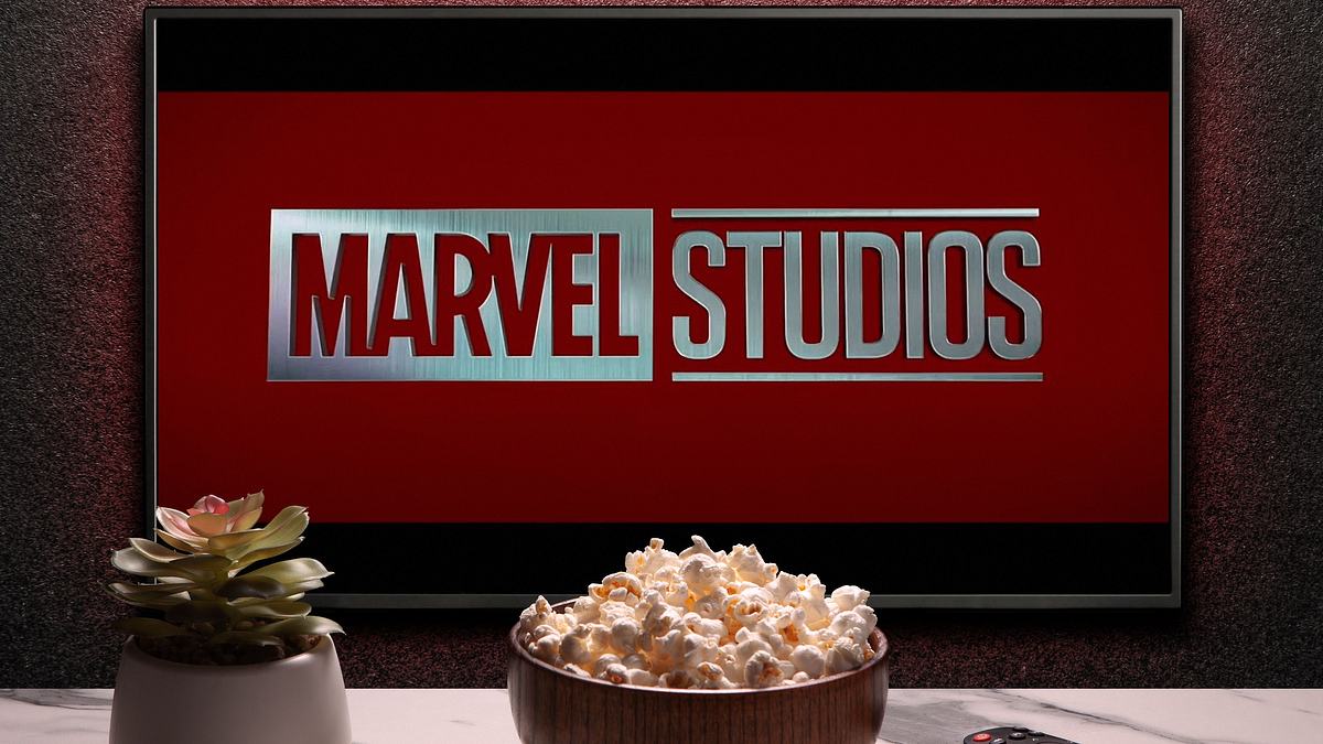 Man sieht einen Fernseher, auf dem „Marvel Studios“ steht. Vor dem Fernseher sieht man eine keine Pflanze, eine Schüssel mit Popcorn und eine Fernbedienung.