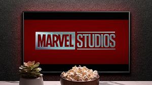 Man sieht einen Fernseher, auf dem „Marvel Studios“ steht. Vor dem Fernseher sieht man eine keine Pflanze, eine Schüssel mit Popcorn und eine Fernbedienung. - Foto:  IMAGO / Pond5 Images