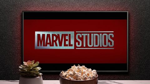 Man sieht einen Fernseher, auf dem „Marvel Studios“ steht. Vor dem Fernseher sieht man eine keine Pflanze, eine Schüssel mit Popcorn und eine Fernbedienung. - Foto:  IMAGO / Pond5 Images